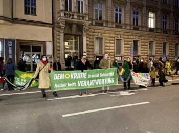 grüne Mitglieder stehen in der Solidarischen Menschenkette in der Luitpoldstraße und halten ein grünes Banner mit der Aufschrift "Demokratie braucht Solidarität, Freiheit heißt Verantwortung"