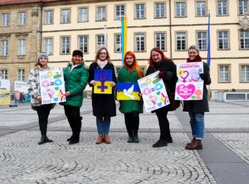 sechs grüne Frauen stehen nebeneinander auf dem Maxplatz in Bamberg und halten Schilder mit feministischen Botschaften in die Kamera